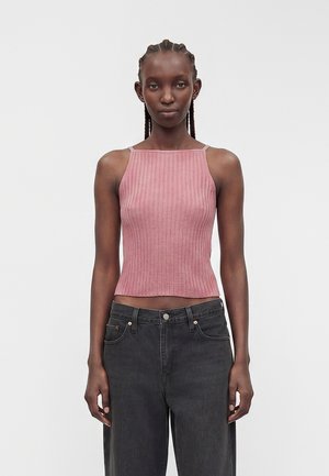 Femme aux cheveux tressés portant un haut rose côtelé sans manches et un jean noir taille haute, debout devant un fond clair uni.
