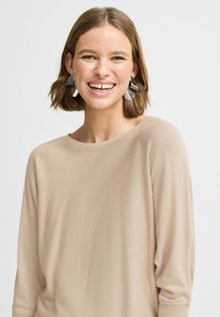 Pull beige à manches longues avec un col rond, fabriqué en tissu doux. Le modèle porte des boucles d'oreilles géométriques argentées et a les cheveux jusqu'aux épaules.