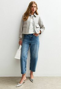 Grijze cropped jack, witte aansluitende top, lichtblauwe high-waisted jeans, zilveren hakken en een kleine witte handtas. Glad textuur.