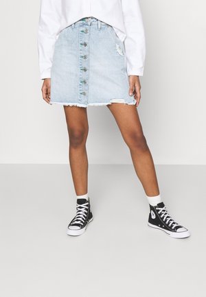 Personne portant une jupe en jean bleu clair à boutons avec un effet usé, une chemise blanche à manches longues, des chaussettes blanches et des baskets montantes noires.