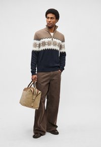 Suéter de malha com padrão azul-marinho e creme, com gola de meio fecho éclair, combinado com calças largas de bombazine castanhas e um saco tipo duffle bege e castanho.