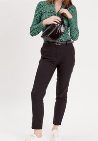 Femme portant un chemisier vert à motifs et un pantalon noir, tenant un sac bandoulière en cuir noir, debout en baskets blanches sur un fond blanc.