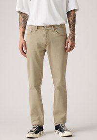Jeans de mezclilla beige con un corte recto, cierre de botón y cremallera, cinco bolsillos y costuras en contraste. Combinados con zapatillas de lona negras.