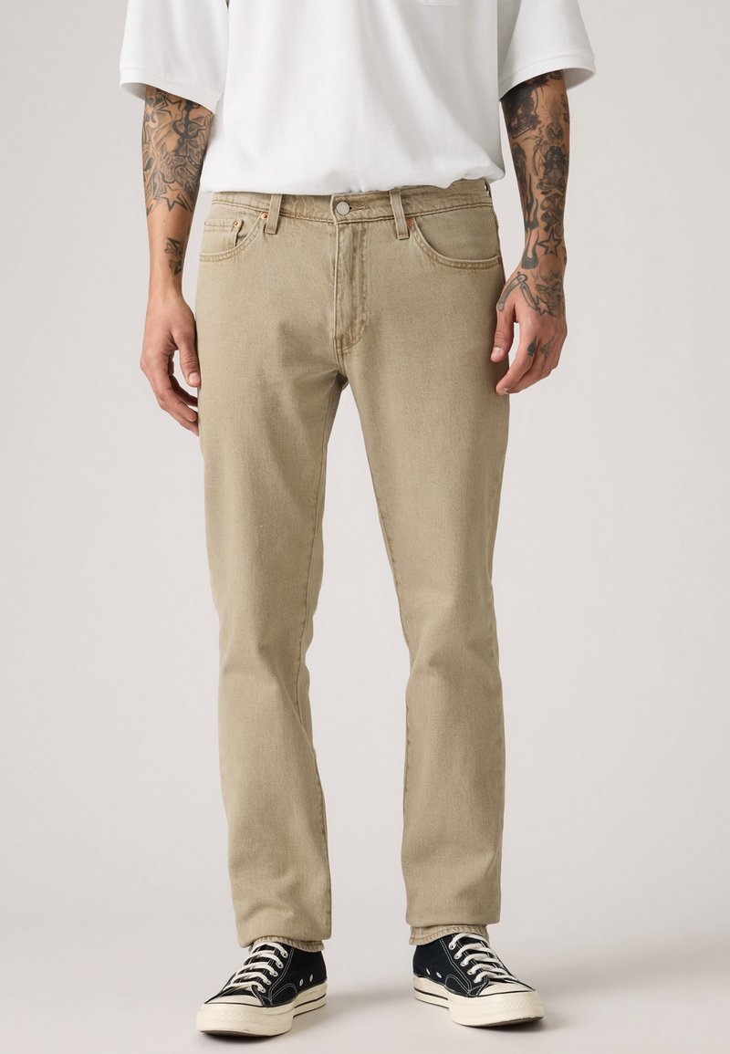 Jeans de mezclilla beige con un corte recto, cierre de botón y cremallera, cinco bolsillos y costuras en contraste. Combinados con zapatillas de lona negras.