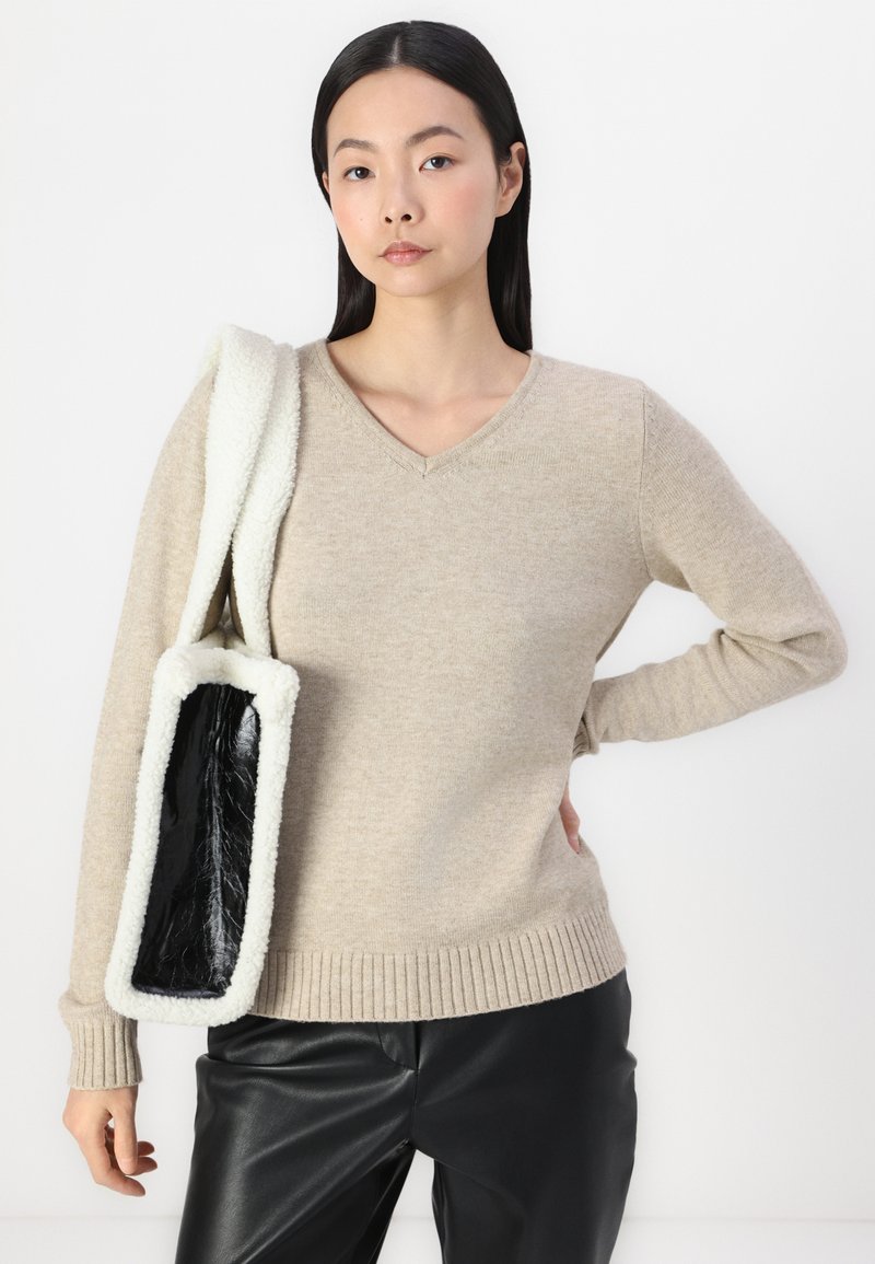 Pull beige à col en V avec un ourlet côtelé et des manches longues, associé à un sac à épaule texturé noir et blanc. Pantalons en cuir noirs.