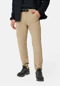 Beige, leichte Hose mit schmaler Passform, elastischem Bund und Gürtelschlaufen; kombiniert mit schwarzen Wanderschuhen mit gelben Akzenten.