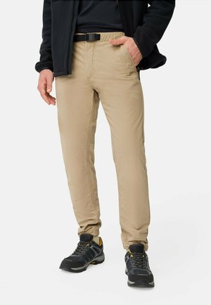 Beige, leichte Hose mit schmaler Passform, elastischem Bund und Gürtelschlaufen; kombiniert mit schwarzen Wanderschuhen mit gelben Akzenten.