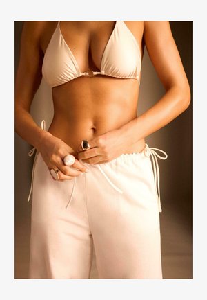 Donna che indossa un top bikini beige e pantaloni chiari, mentre stringe il laccio, con più anelli visibili sulle dita, su uno sfondo neutro.