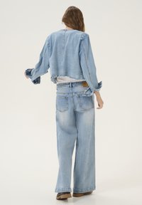 Jachetă din denim tăiată și blugi cu picioare largi în albastru deschis. Margini desfăcute, mâneci lungi cu manșete răsucite și etichetă simplă pe spate.