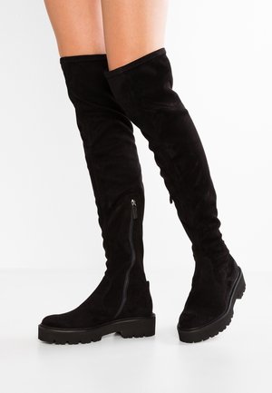 Une personne porte des bottes noires au-dessus du genou, dotées de fermetures éclair latérales et de semelles crantées épaisses, sur fond blanc.