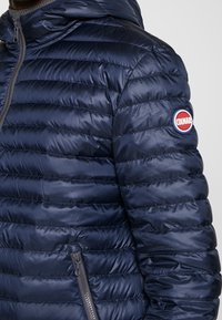 Marinblå pufferjacka med huva, med quiltad yta, grå dragkedjor och en cirkulär Colmar-logotyp på vänster ärm.