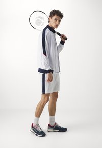 Sportoutfit für Herren, bestehend aus einer weißen Zip-Jacke mit marineblauen und roten Akzenten, weißen Shorts und blauen Sneakers mit pinken Details.