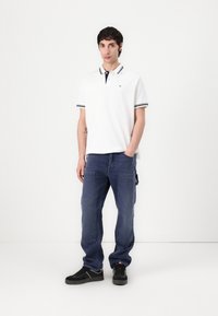 Bílá polo tričko s námořnickým límcem a pruhovanými detaily, kombinované s modrými cargo kalhotami. Model nosí černé tenisky. Jednoduchý a ležérní design.