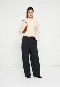 Cremfarbener, gerippter Strickpullover mit Rundhalsausschnitt, kombiniert mit schwarzer, gestreifter weit geschnittenen Hose. Trägt eine kleine braune Lederhandtasche.