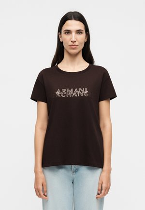 Mujer con cabello largo y oscuro, que lleva una camiseta negra de Armani Exchange y vaqueros azul claro, de pie frente a un fondo blanco liso.