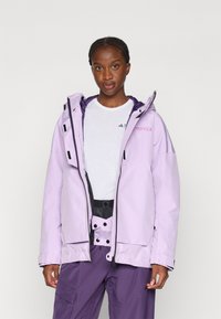 Giacca impermeabile viola con cappuccio, cerniere nere, tasche frontali e dettaglio del logo. Indossata sopra una camicia bianca e pantaloni viola.