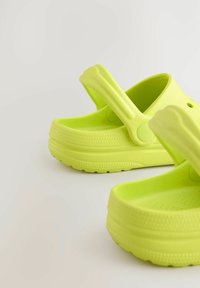 Fényes lime zöld klumpák sima felülettel, nyitott háttal és széles pánttal. A talp mintázott tapadós felülettel rendelkezik, amely biztosítja a jó tapadást.