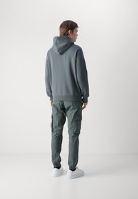 Kapucar zelenega puloverja, v kombinaciji z usklajenimi cargo hlačami s stranskimi žepi. Bele superge dopolnjujejo outfita.