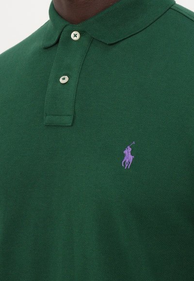 Zaļš polo krekls ar teksturētu audumu, ar apkakles kaklu, divām baltām pogām un purpura logotipa akcentu kreisajā krūtī.