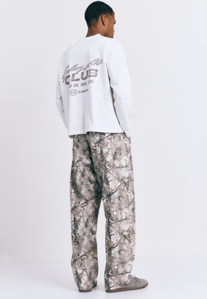 Muž v bílé košili s dlouhým rukávem a černým nápisem, volných kalhotách s maskáčovým vzorem, stojící bokem s rukama v kapsách.