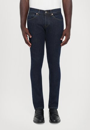Jeans de denim azul oscuro con un corte ajustado, diseño de cinco bolsillos y detalles de costura sutiles. Combinados con zapatos negros.