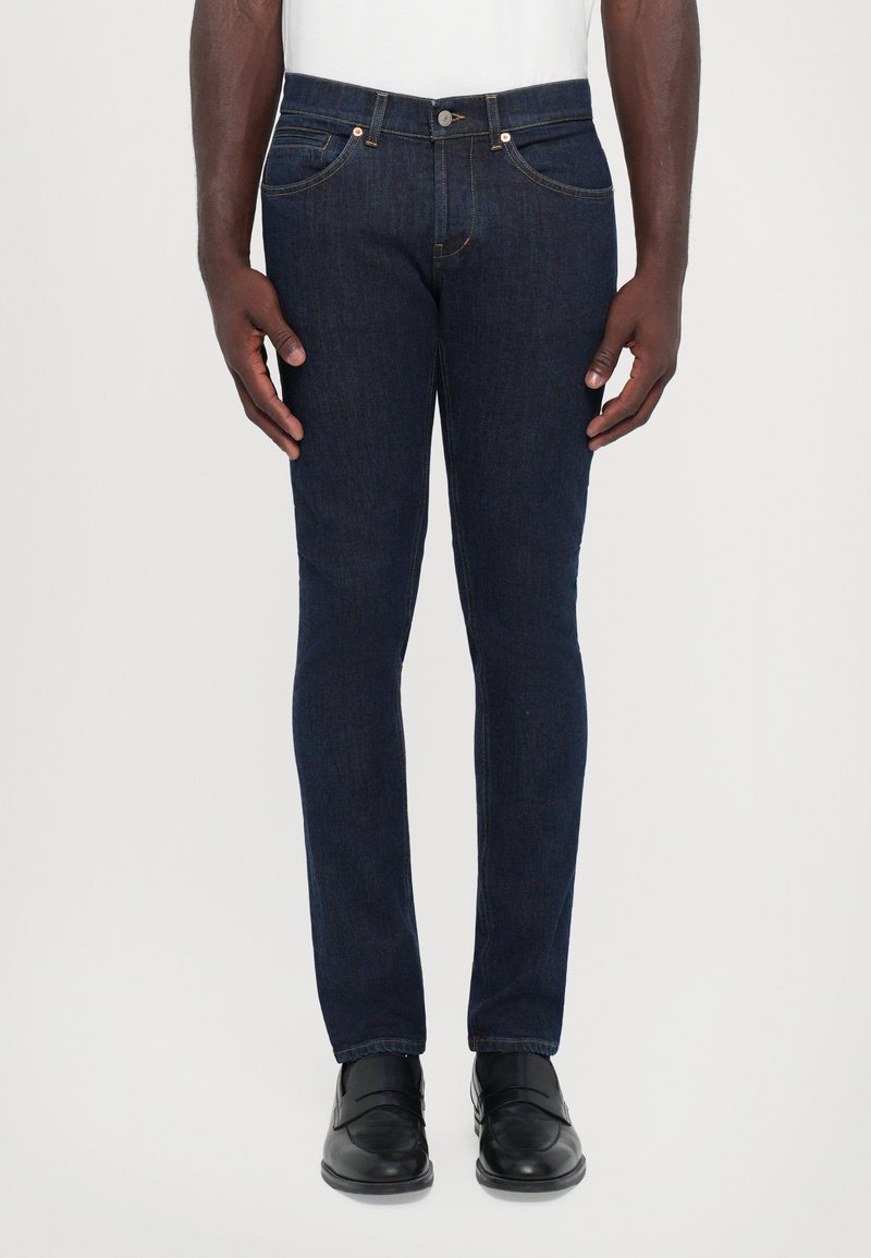 Jeans de denim azul oscuro con un corte ajustado, diseño de cinco bolsillos y detalles de costura sutiles. Combinados con zapatos negros.