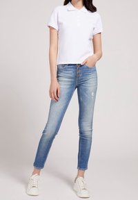 Camisa polo blanca de manga corta, de diseño ajustado, combinada con jeans skinny de tiro medio en un tono azul claro que presentan un cierre de cuatro botones marrones y un ligero desgaste.