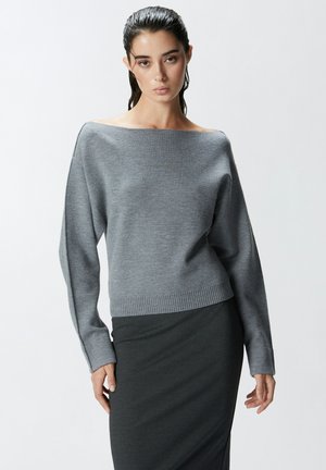 Maglione grigio lavorato a maglia con scollatura off-shoulder, maniche lunghe e orlo a coste, abbinato a una gonna fitted grigio scuro. Texture liscia.
