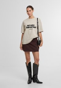 Camiseta beige de gran tamaño con texto negro, combinada con una falda de punto marrón y botas de cuero negras altas con punta afilada y diseño arrugado.