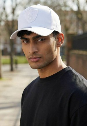 Junger Mann, der eine weiße Raiders-Kappe und ein schwarzes T-Shirt trägt, steht im Freien vor unscharfem Hintergrund mit Bäumen und Gebäuden.