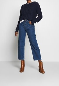 Laivastonsininen palmikkoneule, jossa pitkät hihat, puettuna vaalean yläosan päälle; yhdistetty korkeavyötäröisiin, leveälahkeisiin sinisiin farkkuihin ja ruskeisiin korollisiin saappaisiin.