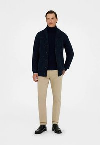 Marineblauwe gebreide cardigan met een gestructureerd patroon, grote knopen, gedragen over een donker coltrui. Gecombineerd met beige broek en zwarte schoenen.