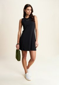 Abito nero senza maniche con scollatura rotonda, caratterizzato da una texture sottile e un dettaglio con logo. Abbinato a sneakers bianche e con una borsa verde.