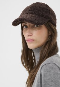 Brown fleece cap with a curved brim, textured surface, and stitched panels. Model wears a gray turtleneck sweater.