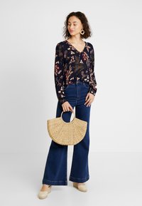Mörk topp med blommönster och långa ärmar, kombinerad med hög midja denim flared byxor. Håller en rund vävd stråväska.