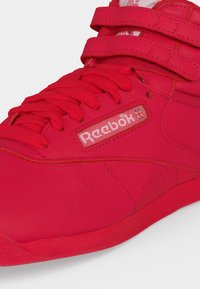 Κόκκινο παπούτσι Reebok με κορδόνια, δύο λουριά με βέλκρο και σημαία Union Jack στο λογότυπο στην πλαϊνή πλευρά σε λευκό φόντο.