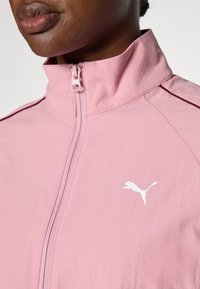 Rosa lättviktsjacka med hög krage, frontdragkedja och vit Puma-logotyp. Har ett slätt tyg och subtila sömdetaljer.