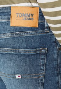 Lähikuva sinisistä farkuista, joissa on takatasku, jossa on pieni Tommy Hilfiger -lipputunnus ja vaaleanruskea nahkalaatta, jossa lukee "Tommy Jeans".
