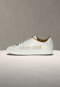 Baskets basses en cuir blanc avec broderie du logo "roberto cavalli" beige et lacets blancs sur un fond neutre.