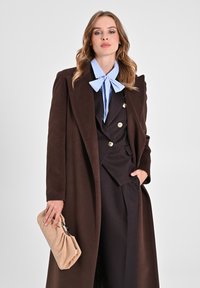 Un lungo cappotto marrone sopra un completo scuro con bottoni dorati, completato da un papillon a righe blu chiaro e una borsa beige. Texture dei tessuti liscia.