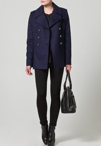Tmavě modrý dvouřadý vlněný kabát s velkými klopami a stříbrnými knoflíky, v kombinaci s černými úzkými kalhotami a černou kabelkou.