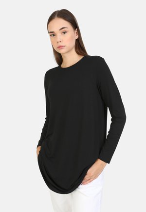 Black long-sleeve top made of soft fabric, featuring a rounded neckline and a curved hem. Model wears it with hands in pockets and white pants.