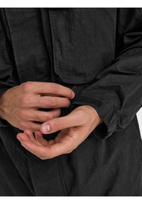 Palton negru dintr-un material texturat. Se văd mâini care ajustează închiderea cu arici de la manșetă. Include un detaliu cu buzunar pe piept și manșete elasticizate.