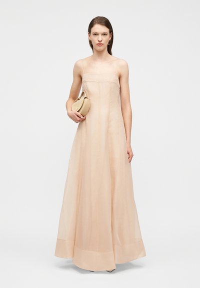 Γυναίκα που στέκεται, φορώντας ένα μακρύ, διάφανο, ροζ-ροδακινί φόρεμα με λεπτές τιράντες, κρατώντας μια μικρή μπεζ τσάντα χειρός με σκουλαρίκια κρίκους.