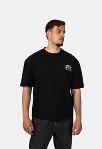 Černé bavlněné tričko s krátkými rukávy a kulatým výstřihem, na levé části hrudi s bílým logem "CRYSTAL". Kombinováno s tmavými džínovými kalhotami.