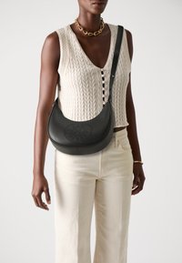 Sac à bandoulière en cuir noir avec une forme incurvée et un design perforé à l'avant. Associé à un haut en tricot crème et à un pantalon de couleur claire.