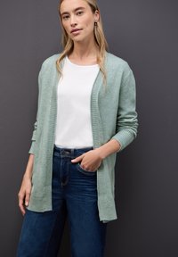 Hellgrüner Cardigan aus weichem Material, mit offenen Vorderteil und gerippten Akzenten, getragen über einem weißen Oberteil und kombiniert mit dunkelblauen Jeans.