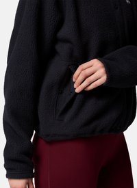 Hand die de drukknoopzak op een zwarte gestructureerde fleecejas aanpast, gedragen over bordeaux legging.