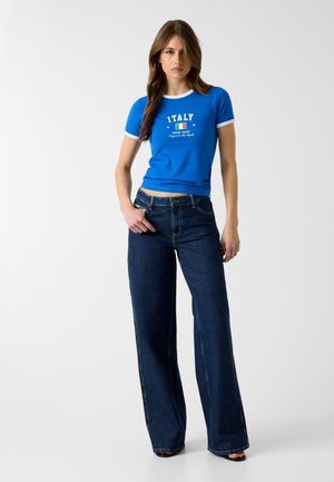 Mujer con camiseta gráfica azul de Italia y vaqueros anchos oscuros de pierna ancha, de pie contra un fondo blanco liso.