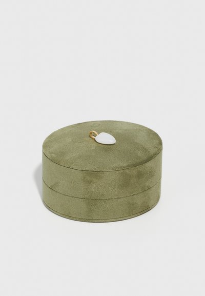 Cutie de bijuterii rotundă, din suede verde măsliniu, având o textură netedă și un accent subțire de aur cu o piatră albă în formă de lacrimă deasupra.